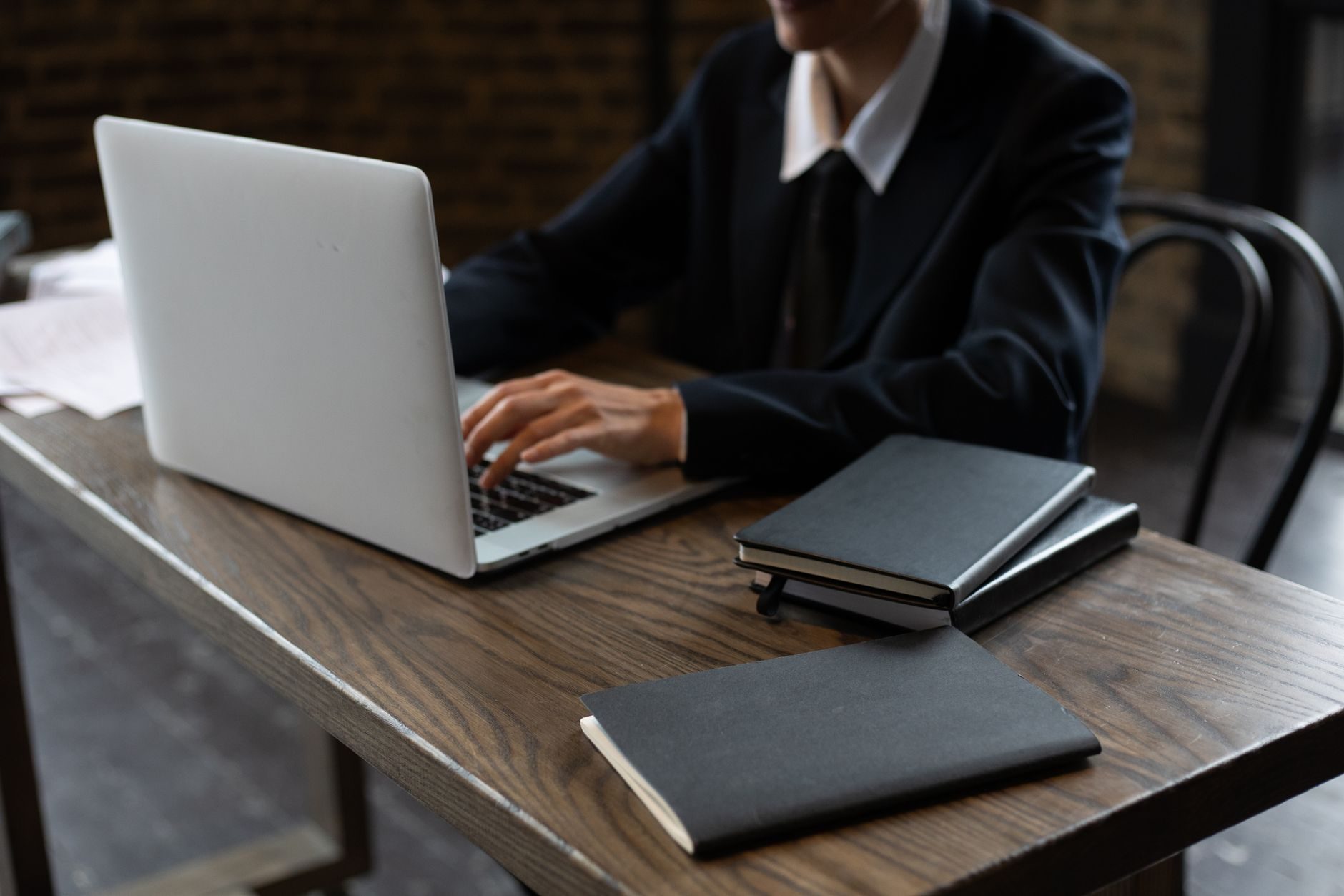 Primer plano de una persona con traje escribiendo en un portátil en un escritorio de madera con cuadernos.